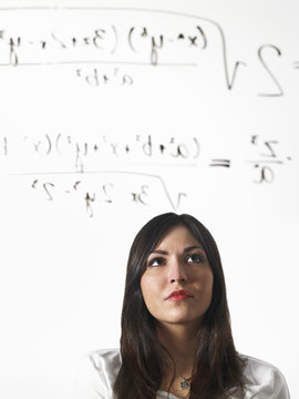 A young woman writing a mathematical equation with black marker on a clear seethrough surface and standing back to consider it.