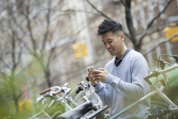 City life in spring. A man in a blue sweater by a row of parked bicycles. Checking his messages on a smart phone.