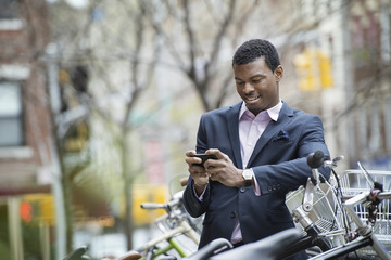 City life in spring. A young man in a blue suit, by a bicycle park. Checking his smart phone for messages.