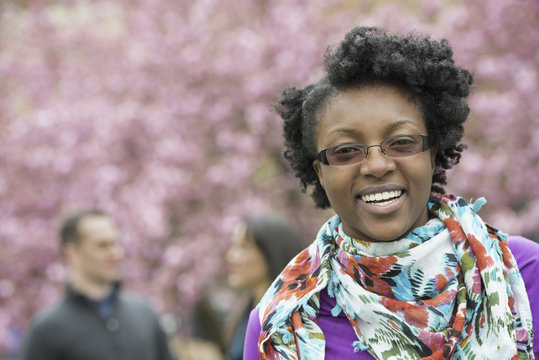 A group of people under the cherry blossom trees in the park. A young woman smiling and looking at the camera. Wearing a floral scarf.