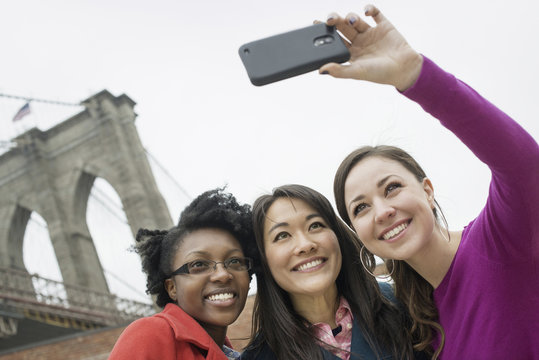 New York city, the Brooklyn Bridge crossing over the East River. Three women in a row, smiling, as one takes a picture with a smart phone.