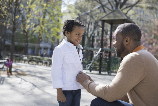 A New York city park in the spring. Sunshine and cherry blossom. A father kneeling and buttoning his son's jacket.