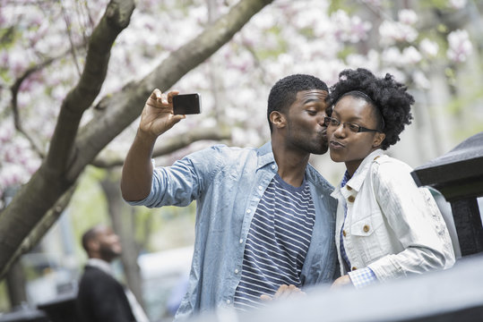 Outdoors in the city in spring. An urban lifestyle. A man kissing a woman and taking a photograph with a handheld mobile phone.