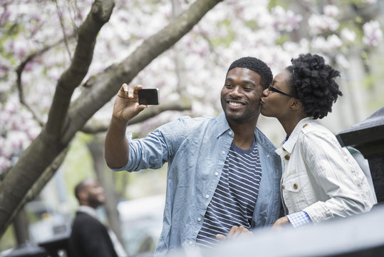Outdoors in the city in spring. An urban lifestyle. A woman kissing a man and taking a photograph with a handheld mobile phone.