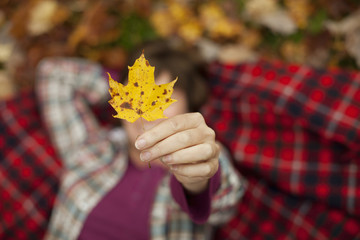 A woman lying on a red tartan picnic blanket, looking upwards, holding a maple leaf. 