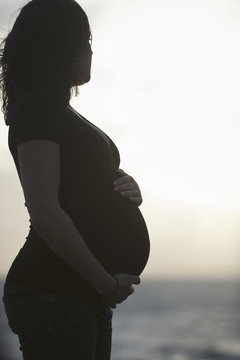 Silhouette of a heavily pregnant woman standing on a beach, with one hand resting on the top of her belly and one supporting it.