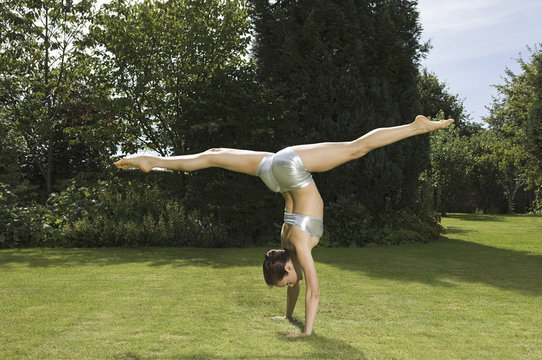 A young woman doing a splits handstand on the grass in a garden. 