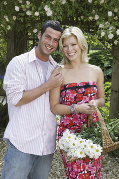 Two people, a  smiling couple, in a garden. A young woman carrying a trug of cut flowers.