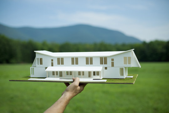 A rural scene and mountain range, and a person holding a scale model of a new building.