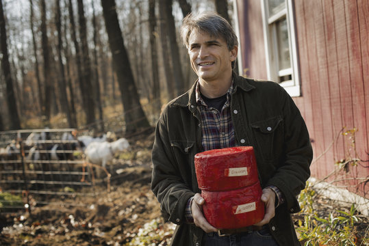 A man standing in a farm courtyard, holding two blocks of goat's cheese.