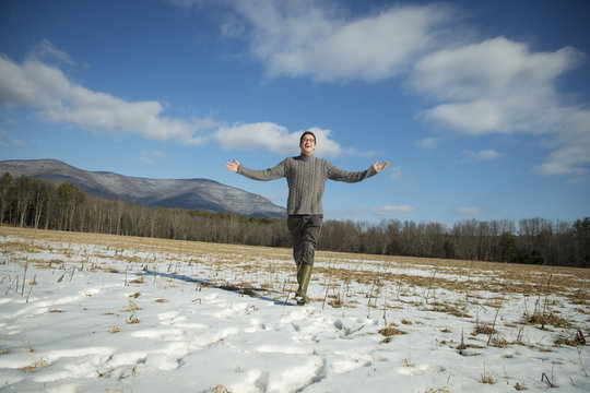 A man in a cable knit jumper and muck boots standing with his arms stretched out, in a snowy rural landscape. 