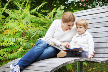 Little boy and his mother sitting on bench in park and reading b