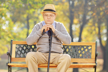 Fototapeta premium Thoughtful senior man with a cane sitting on bench in a park