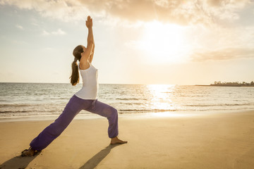 Fototapeta na wymiar Young woman practicing yoga warrior pose on the beach at sunset