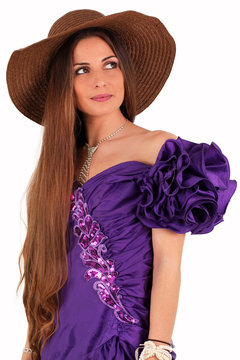 Modebewusste, elegante Frau mit Hut, wundersch&ouml;nem lila Kleid, ausdrucksvollem Make-up und langen, nat&uuml;rlichen Haaren. Das Sch&ouml;nheitsmodel l&auml;chelt und blickt seitlich nach oben.