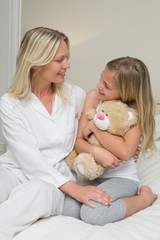 Girl embracing teddy bear while looking at mother in bed