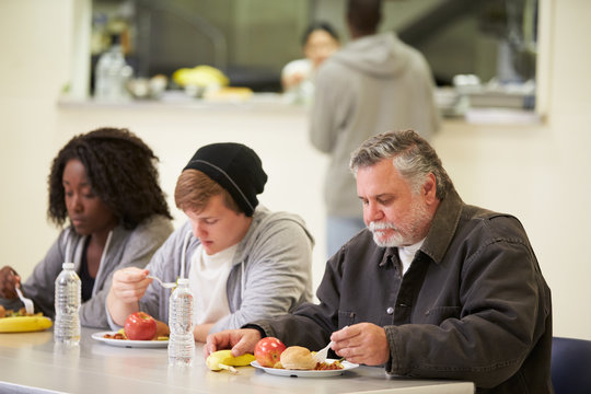 People Sitting At Table Eating Food In Homeless Shelter