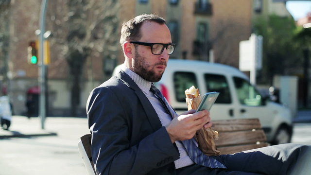 Businessman using cellphone eating baguette on street bench, ste