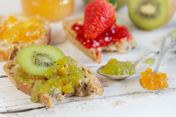 kiwi, orange and strwberry jam with slices of bread on wooden ta