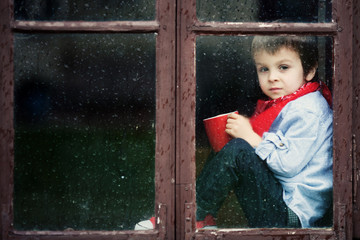 Little boy on the window, drinking tea and watching the rain