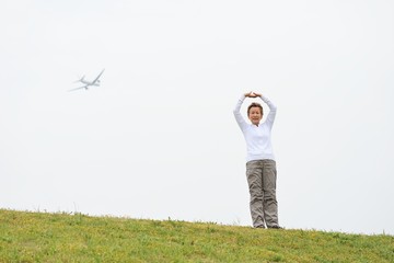 白い空をバックに芝生の上でストレッチ運動をする高齢の日本人女性