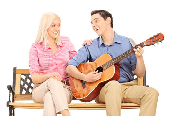 Man playing guitar for a girl seated on bench