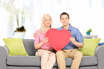 Obraz premium Young couple sitting at sofa and holding big red heart