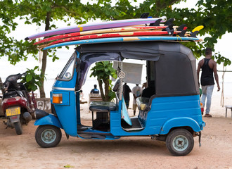 Blue tuk tuk vehicle transporting surfboards on the roof.