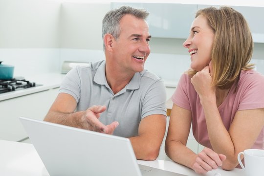Cheerful couple in conversation while using laptop in kitchen