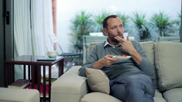 Man eating sandwich and drinking tea while sitting on sofa 