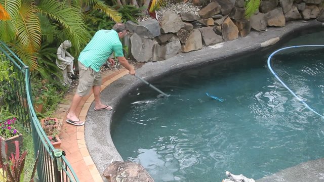 Man cleans pool with long scraper and automatic pool cleaner.