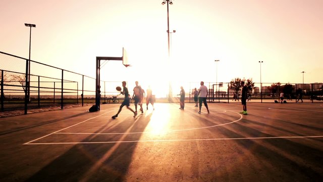 Basketball Match During Sunset.This stock footage shows a group of men playing basketball at sunset. The ground and the sky are enveloped in metallic colors. 