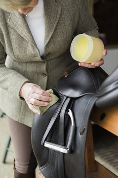 A woman cleaning and preparing tack and saddle, in the courtyard of a riding stable.