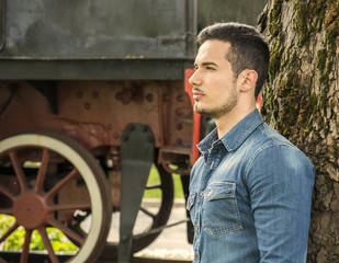 Fototapeta na wymiar Profile of young man in denim shirt near old train, against tree