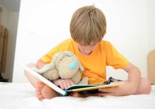little boy reading book with interest lying on bed in his room