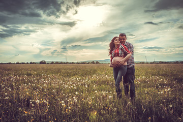 husband and his pregnant wife are walking at sunset in field on
