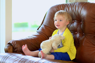 Cute toddler girl sitting on sofa playing with teddy bear