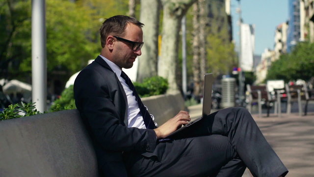 businessman typing on a laptop and sitting on a bench in the str