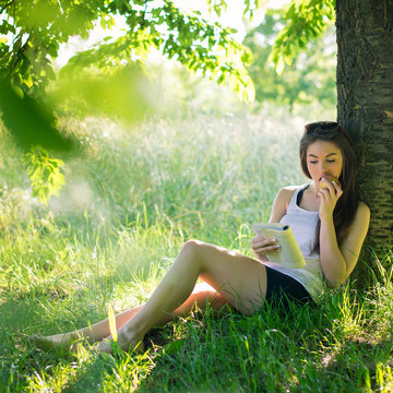 Beautiful young woman reading a book under a tree in the park.