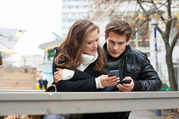 Couple Using Cell Phone While Leaning On Bench