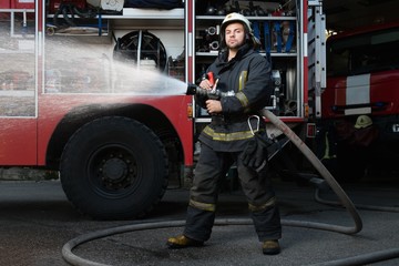Obraz na płótnie Canvas Firefighter holding water hose near truck with equipment