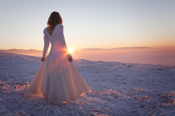 Fototapeta na wymiar Woman in a wedding dress, standing in a winter landscape looking at the sunset on the horizon