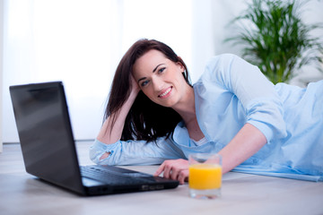 Cheerful young brunette lying floor at home with laptop