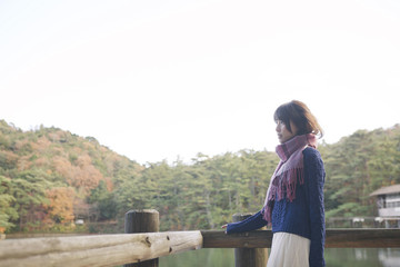 Fototapeta premium 秋の紅葉の公園の池のほとりで池を見つめる女性