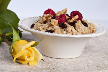 Bowl of freshly made oatmeal with walnuts, raisins, and raspberries next to a yellow rose