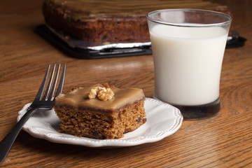 Plate with a square slice of carrot cake topped with caramel frosting and a half a walnut next to a cold glass of 1% milk