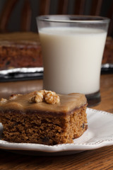 Macro shot of carrot cake topped with a walnut next to a glass of cold milk with whole cake in background