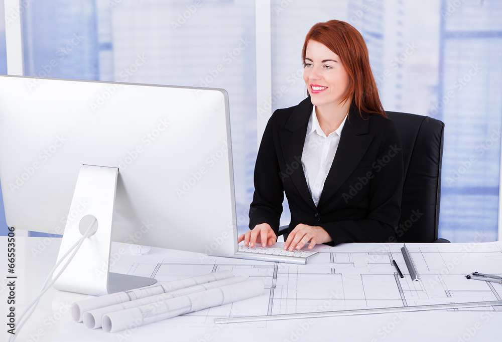 Poster businesswoman using computer with blueprints at desk