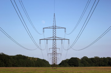 Symmetrical view on a high voltage power line on a rural landscape with forest to both sides. The moon is visible in the background