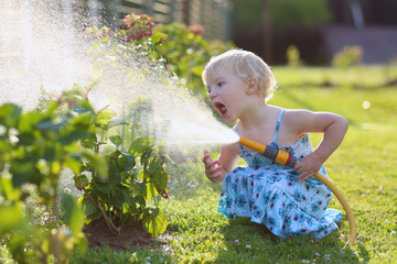 Cute toddler girl watering flowers from hose in summer garden